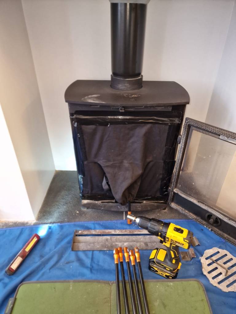 A clean wood-burning stove with a black cover and tools laid out, including a drill and chimney cleaning rods, set on a blue cloth.